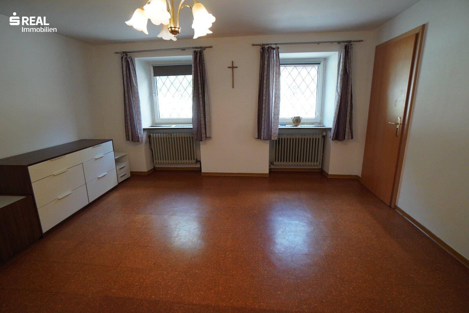 Heller Raum mit zwei Fenstern, Heizkörpern und rotbraunem Bodenbelag, schlicht eingerichtet.