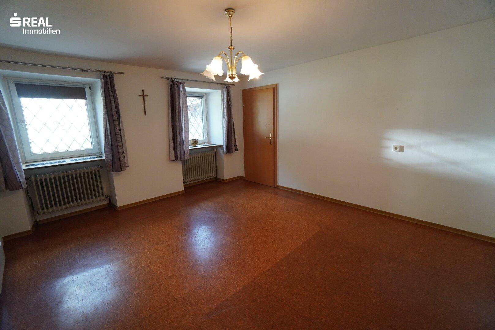 Geräumiger, heller Raum mit zwei Fenstern und rotbraunem Bodenbelag, vielseitig nutzbar.