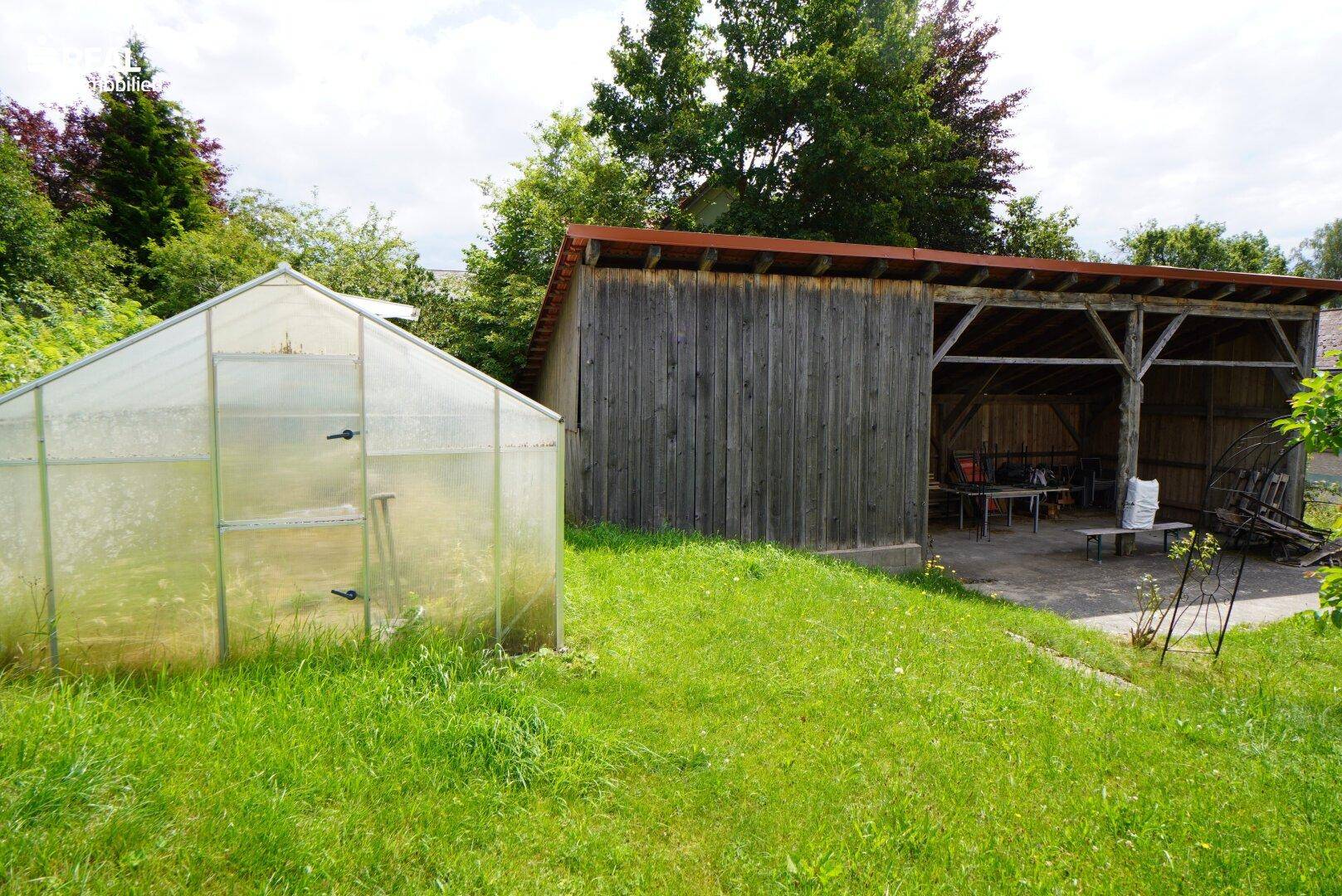 Gewächshaus und überdachter Unterstand in einem grünen Gartenbereich mit Bäumen im Hintergrund.