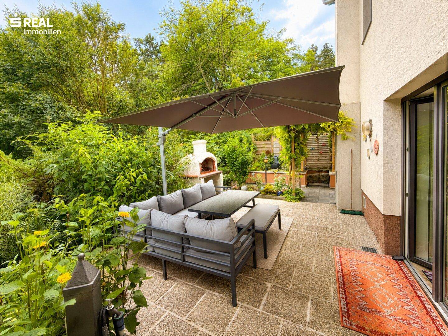 Gemütliche Terrasse mit bequemen Gartenmöbeln, großem Sonnenschirm und gemauertem Ofen im Grünen.