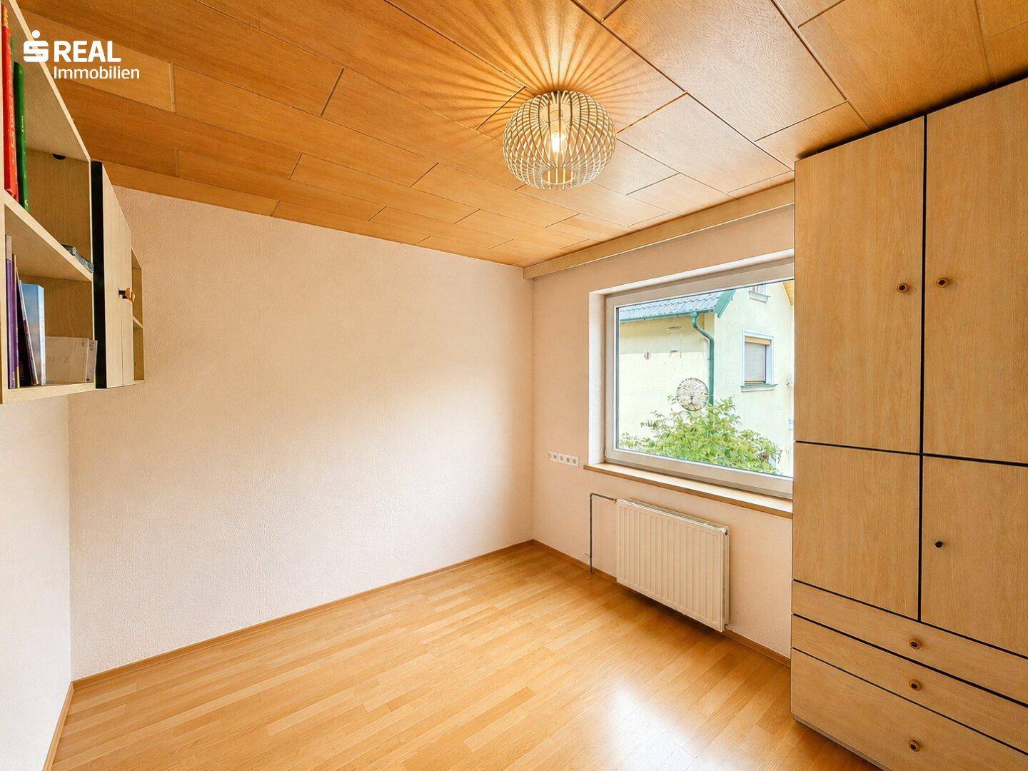 Helles Zimmer mit Holzboden, Holzdecke und einem großen Fenster mit Blick ins Grüne.