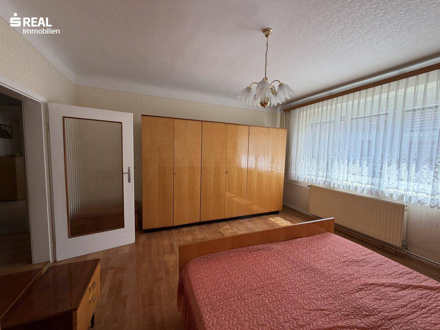 Schlafzimmer mit Holzboden, Einbauschränken und großem Fenster mit Vorhängen.