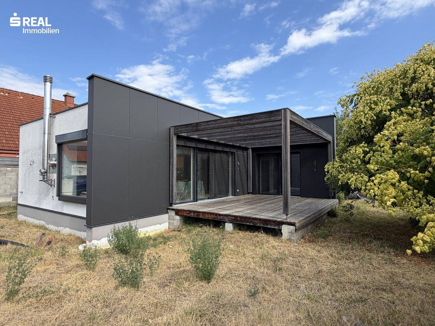 Rückseite des modernen Hauses mit Holzterrasse und Pergola, umgeben von trockener Vegetation.