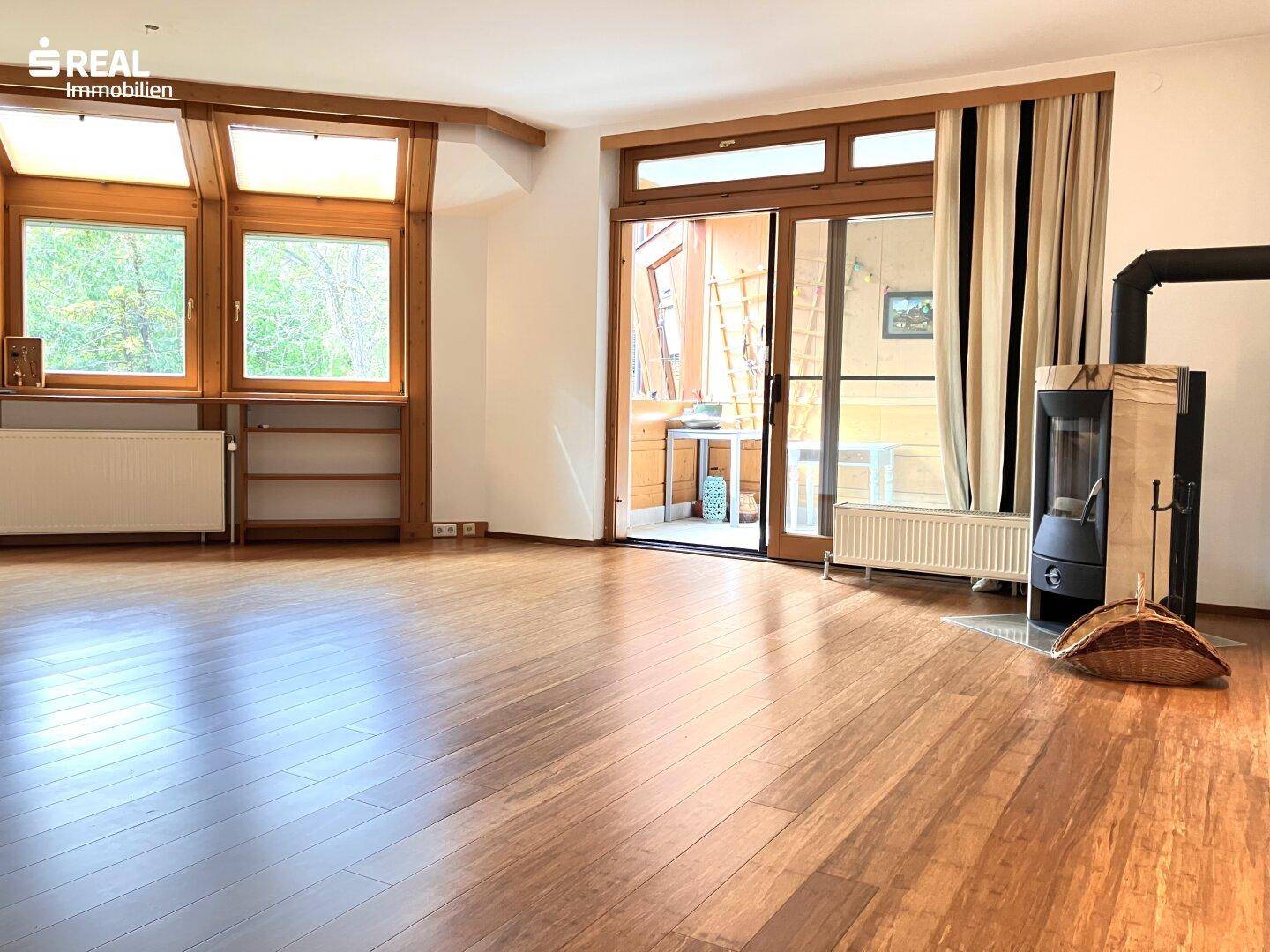 Helles Wohnzimmer mit Holzboden, großen Fenstern, einem modernen Kaminofen und Zugang zum Balkon.