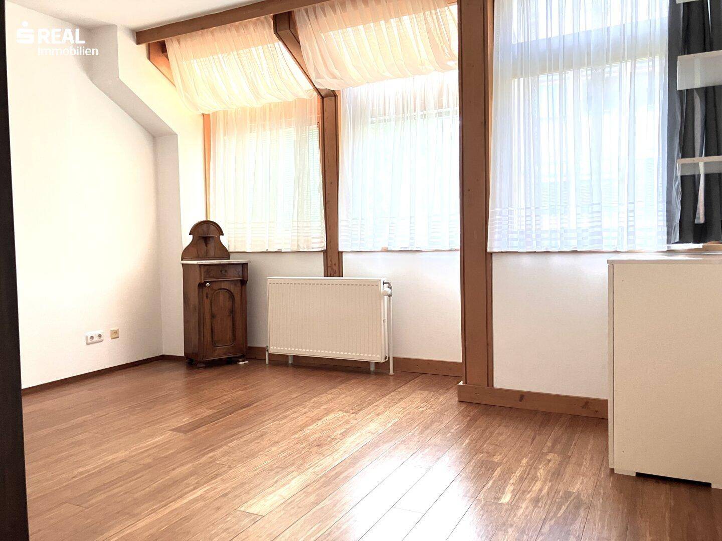 Helles Zimmer mit Holzboden, großen Fenstern und sichtbaren Holzbalken unter der Decke.
