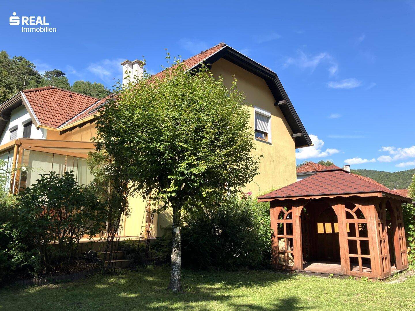 Gelbes Haus mit rotem Ziegeldach, Wintergarten und einem charmanten Holzpavillon im grünen Garten.