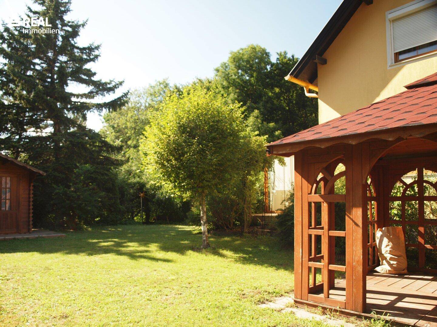 Weitläufiger grüner Garten mit Holzpavillon und altem Baumbestand unter blauem Himmel.