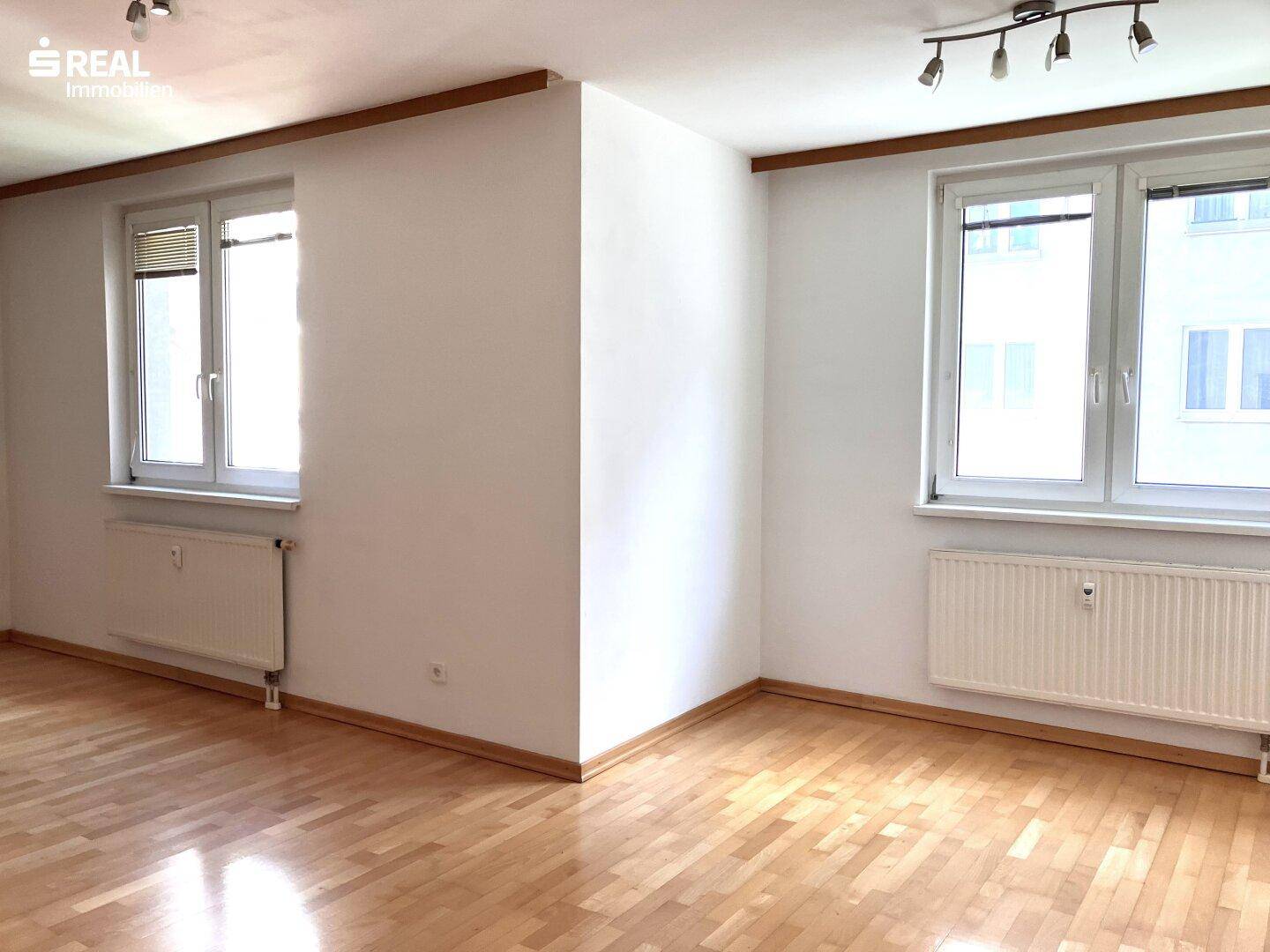 Heller Wohnraum mit zwei großen Fenstern, die viel Tageslicht hereinlassen, und Parkettboden.