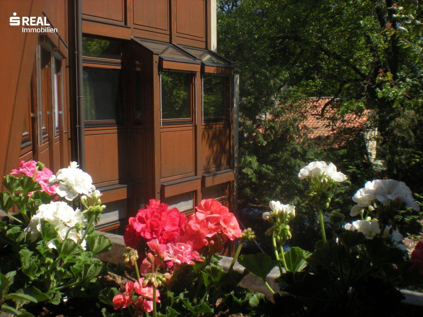 Fassade des Gebäudes mit braunen Fensterrahmen und blühenden Blumen im Vordergrund.