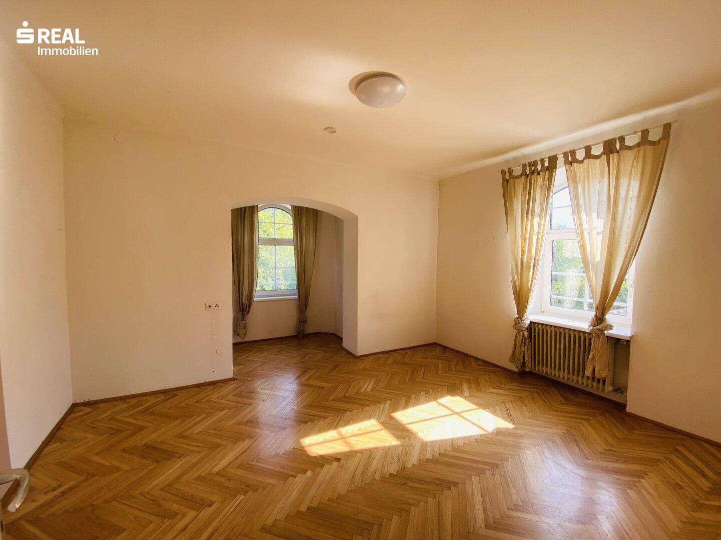 Geräumiges Zimmer mit Fischgrätparkett, zwei Fenstern und viel Tageslicht.