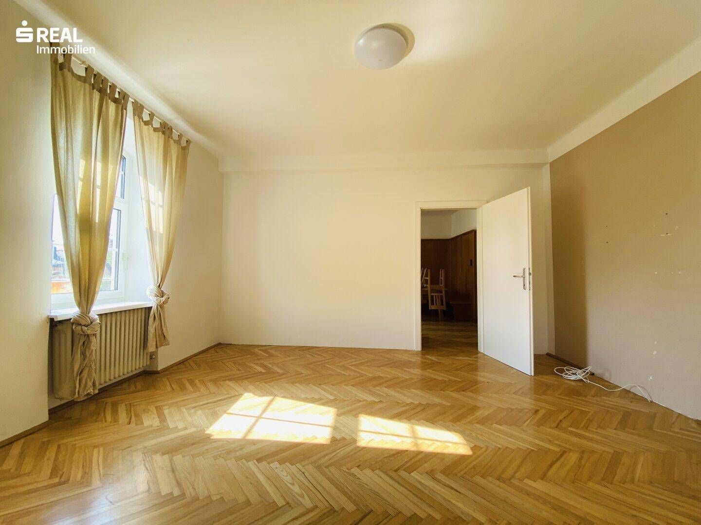 Helles Zimmer mit Fischgrätparkett, hohen Fenstern und Blick ins Grüne.