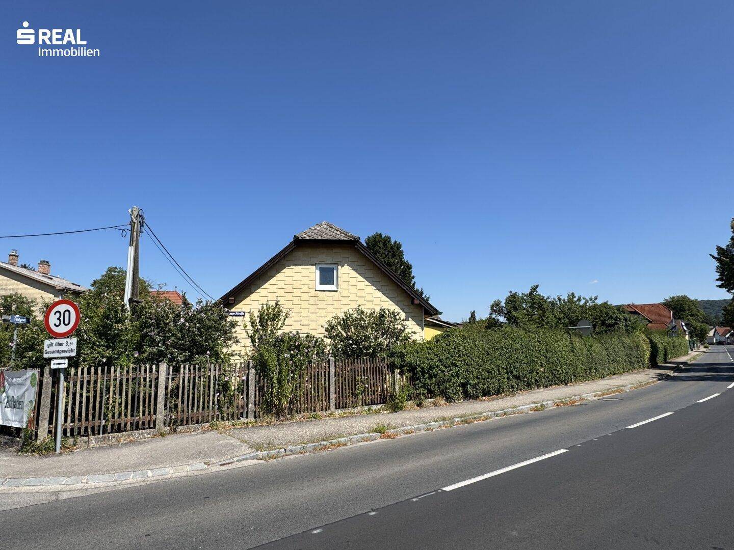 Gelbes Haus mit Holzzaun und Hecke entlang der Straße unter blauem Himmel.