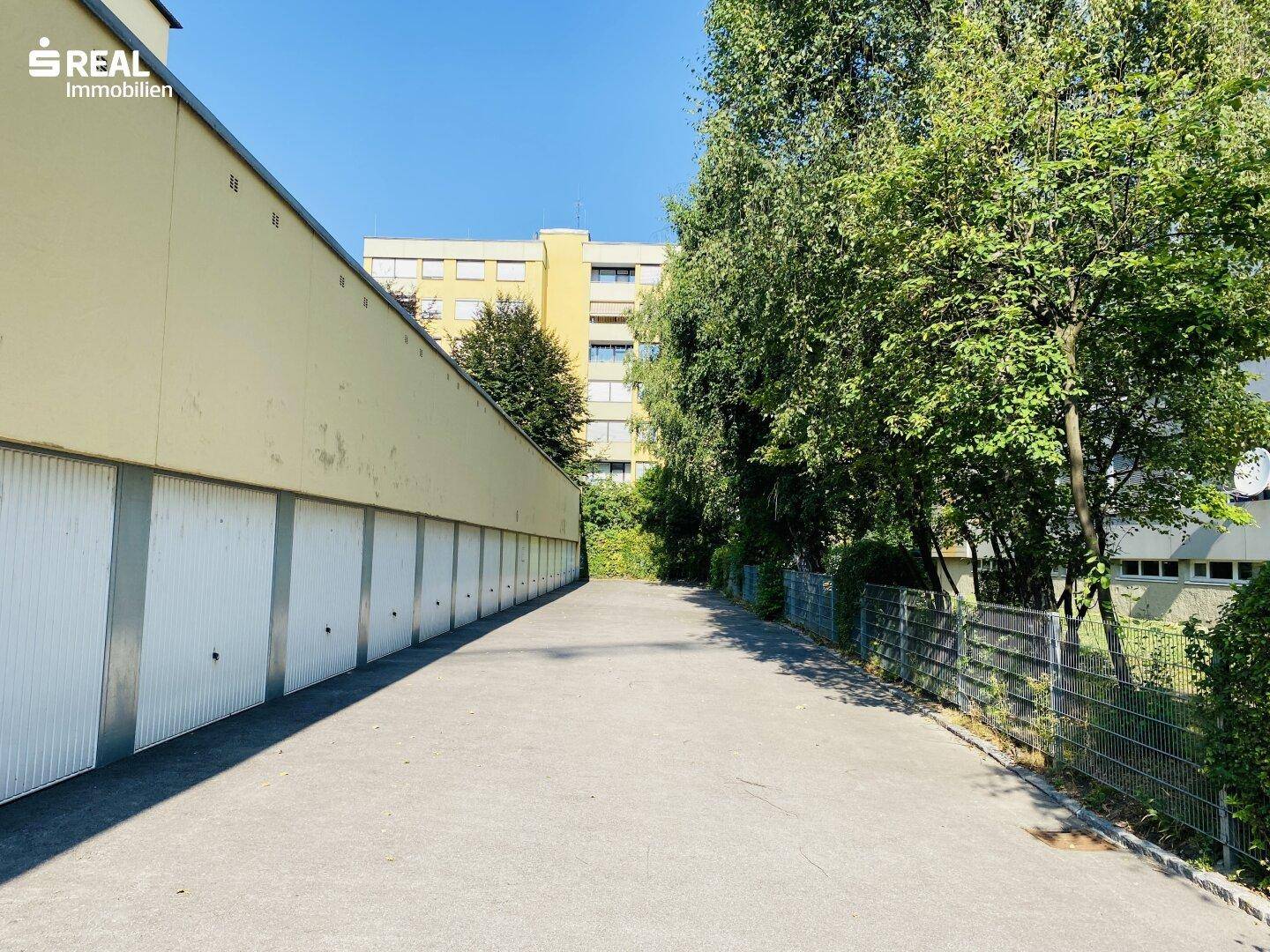 Weitere Ansicht der Garagenreihe mit asphaltiertem Weg und üppigem Grün im Hintergrund.