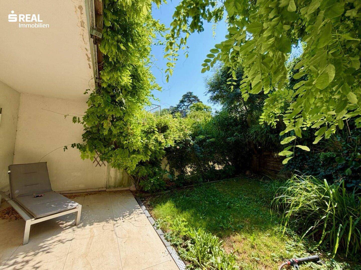 Sonniger Gartenbereich mit gepflasterter Terrasse und Liegestuhl, umgeben von viel Grün.