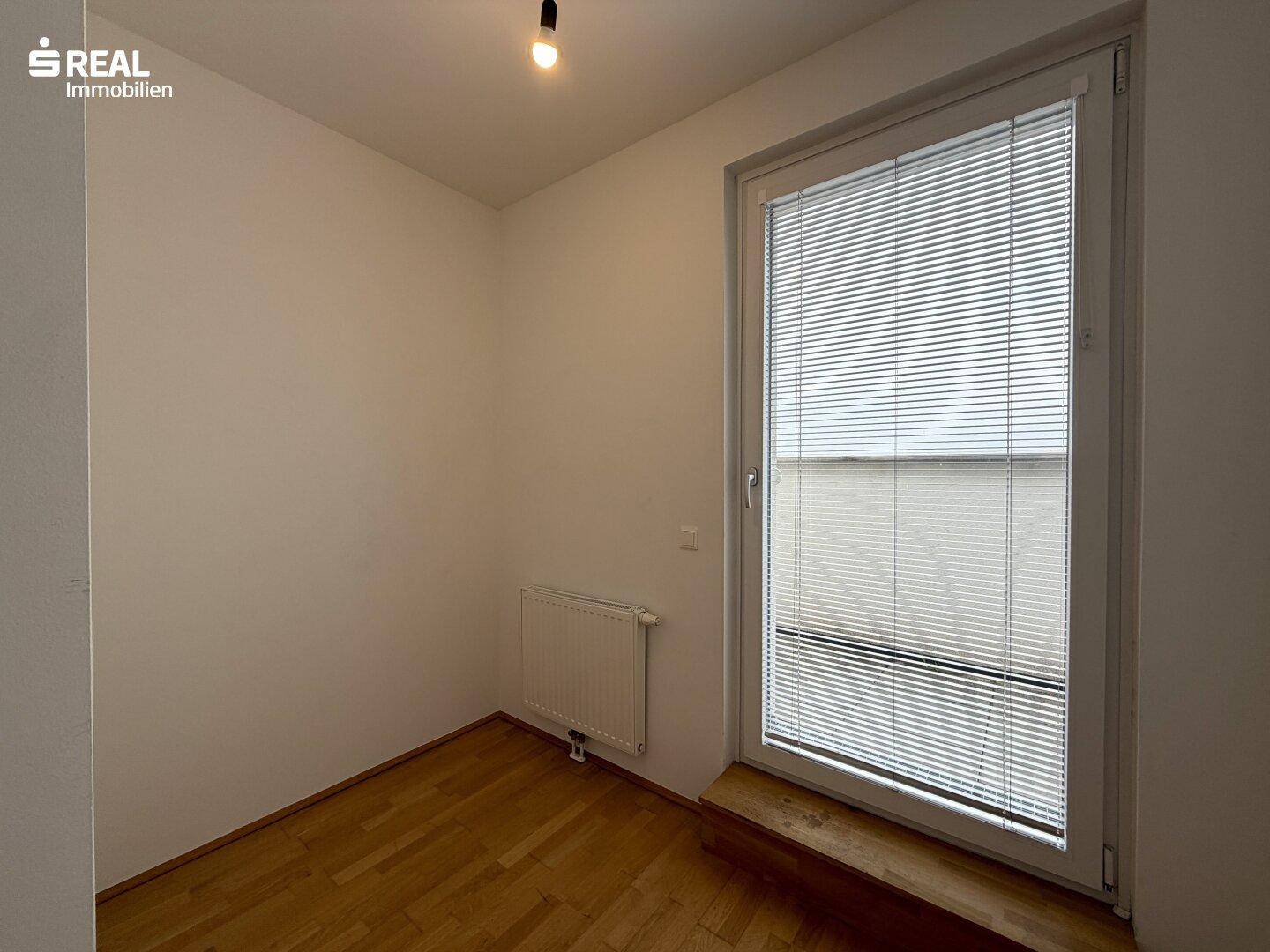 Kleines Zimmer mit Holzboden und einer großen Fenstertür, die viel Licht spendet.