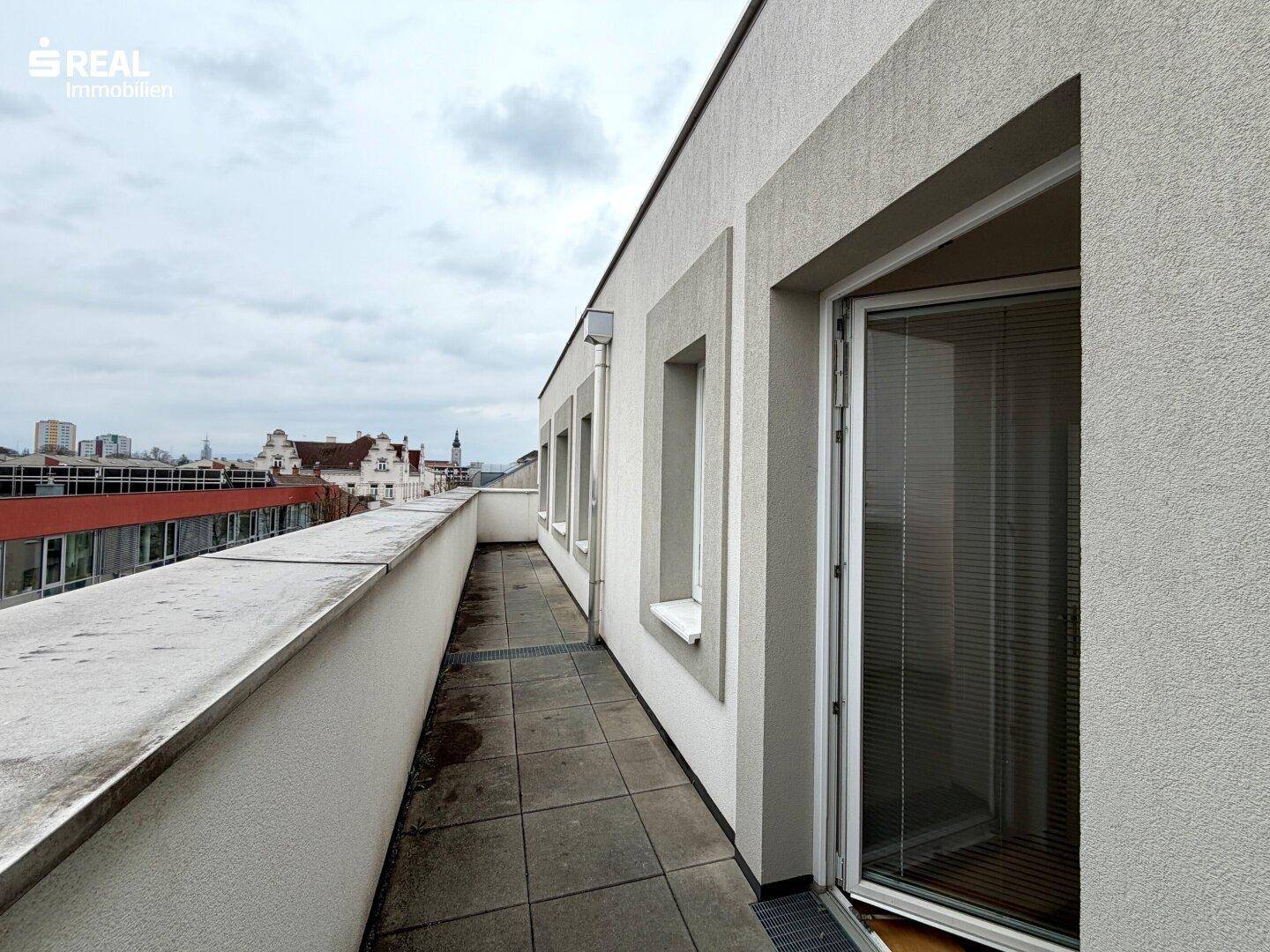 Langer Balkon mit Fliesenboden und Blick auf die umliegenden Gebäude der Stadt.