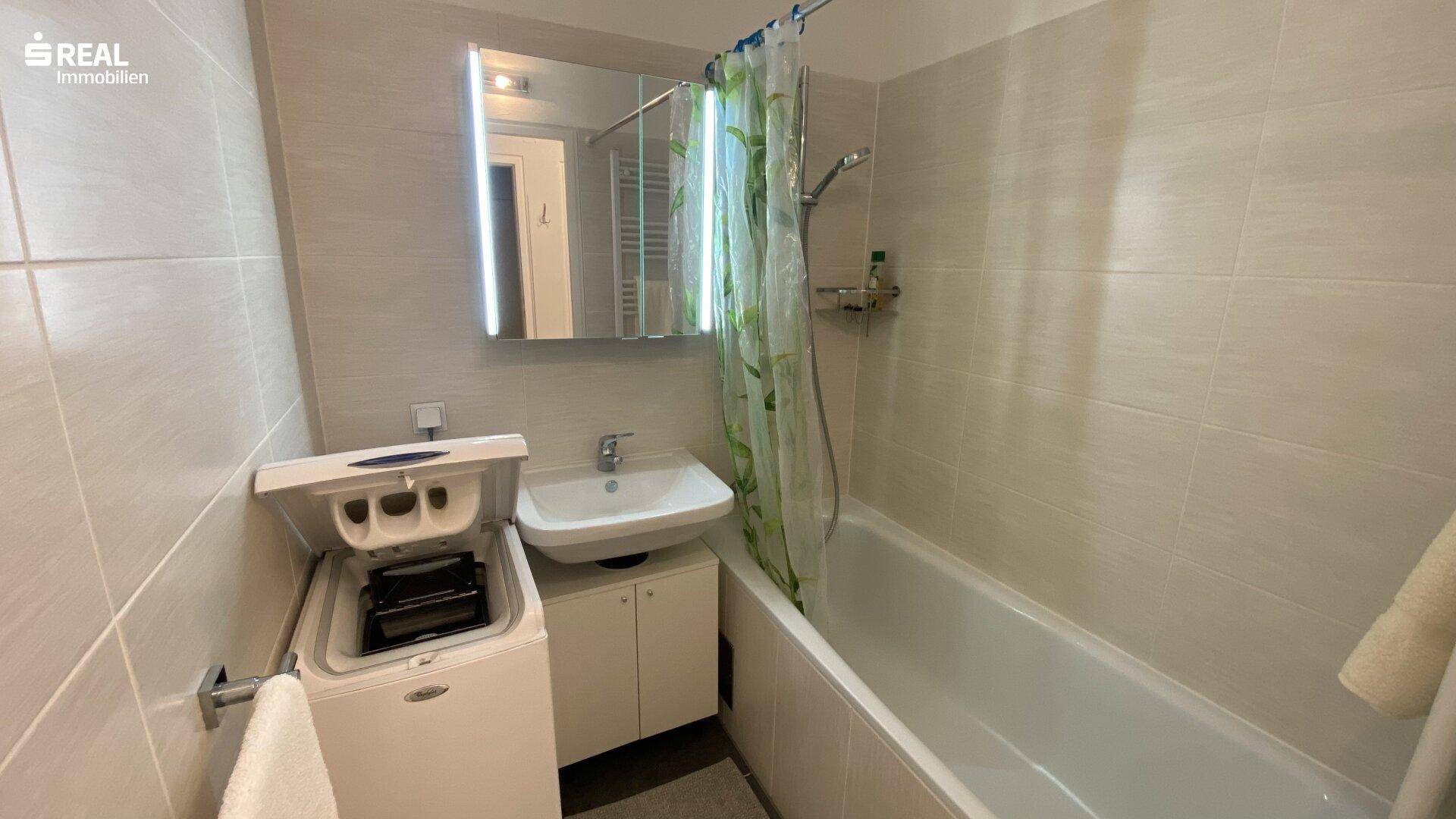 Helles Badezimmer mit Badewanne, Duschvorhang, Waschbecken und Waschmaschine, komplett gefliest.