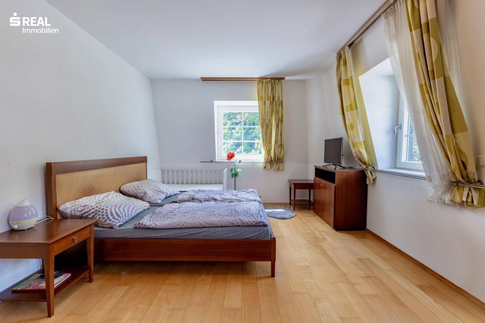 Helles Schlafzimmer mit Holzboden, Doppelbett und Fenstern mit Blick ins Grüne.
