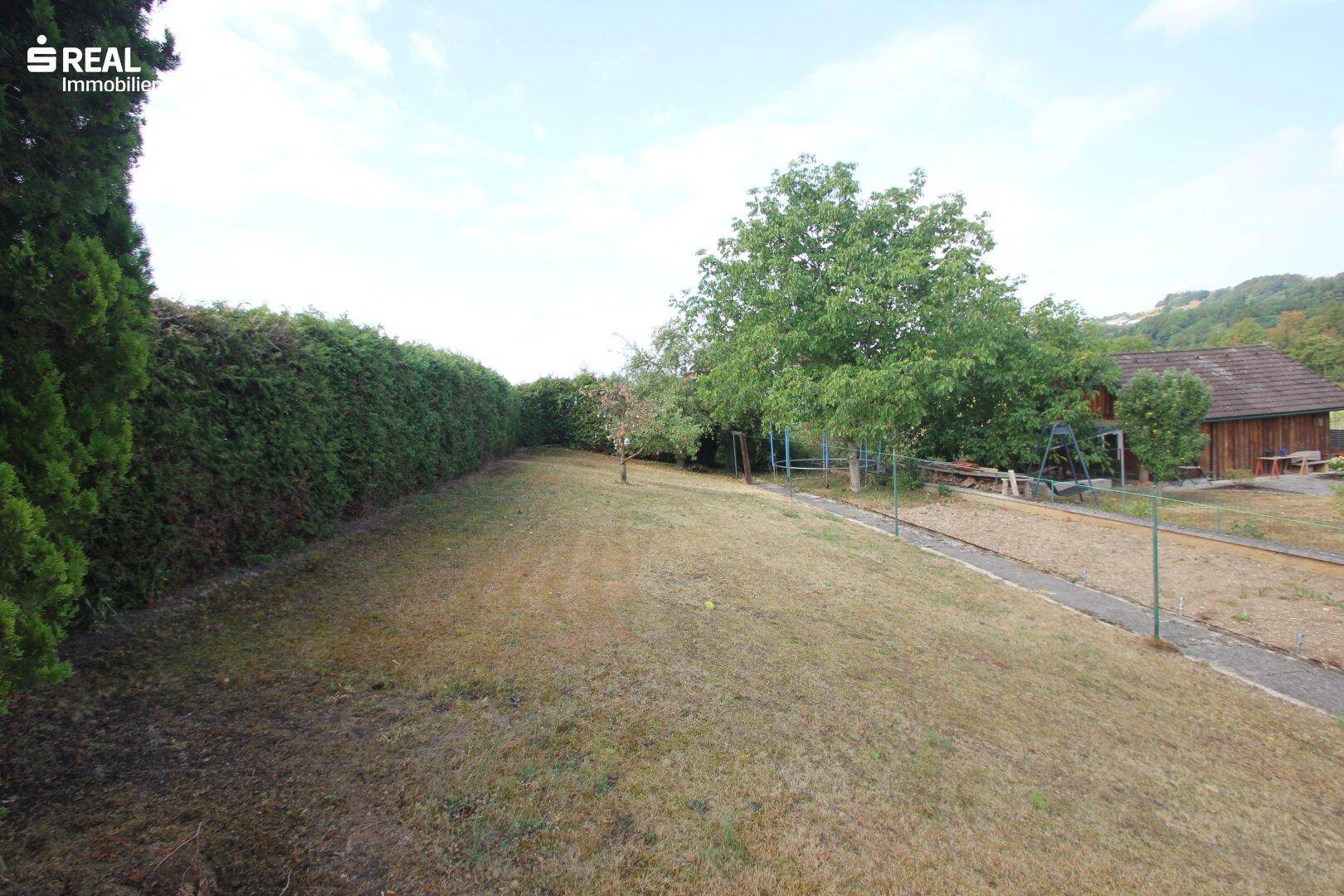 Langer Gartenbereich mit gepflegter Hecke und einigen Bäumen im Hintergrund.