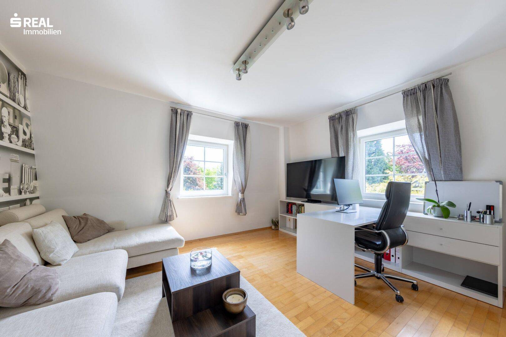 Helles Arbeitszimmer mit Sofa, Schreibtisch und Blick ins Grüne durch die Fenster.