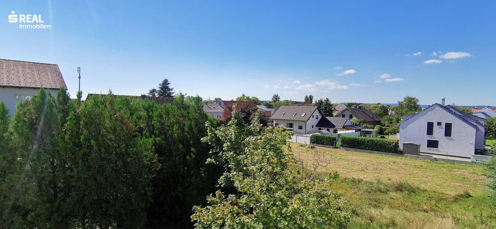 Weitblick über eine ländliche Wohngegend mit Häusern, Bäumen und weiten Feldern unter blauem Himmel.