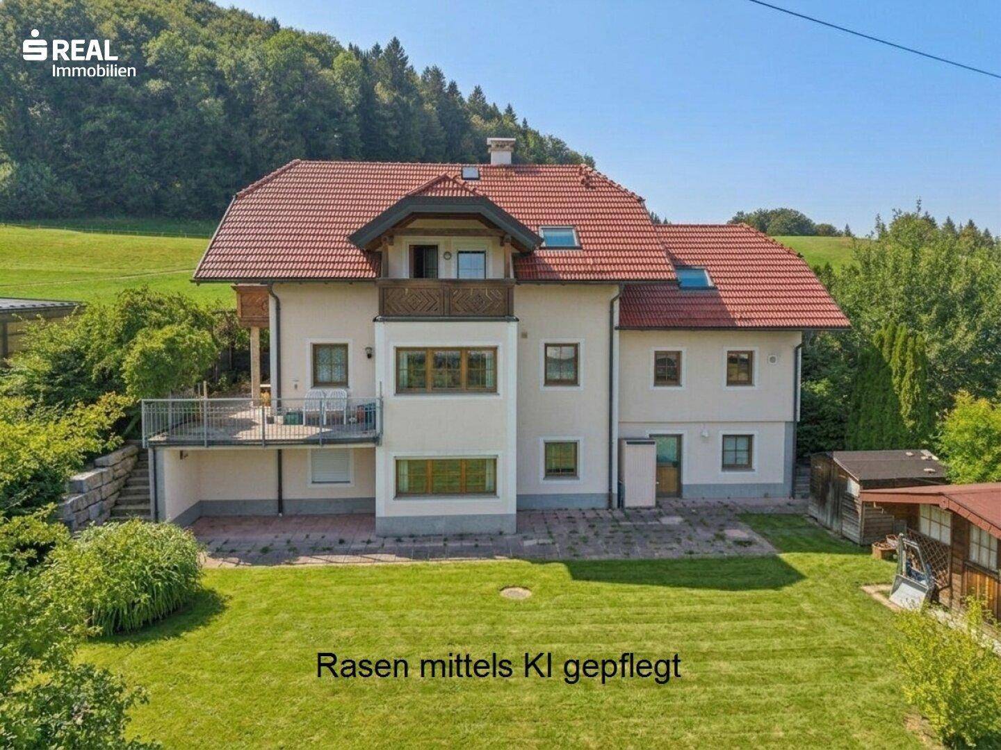 Rückansicht des Hauses mit großer Rasenfläche, Terrasse und Balkon, umgeben von grünen Hügeln.