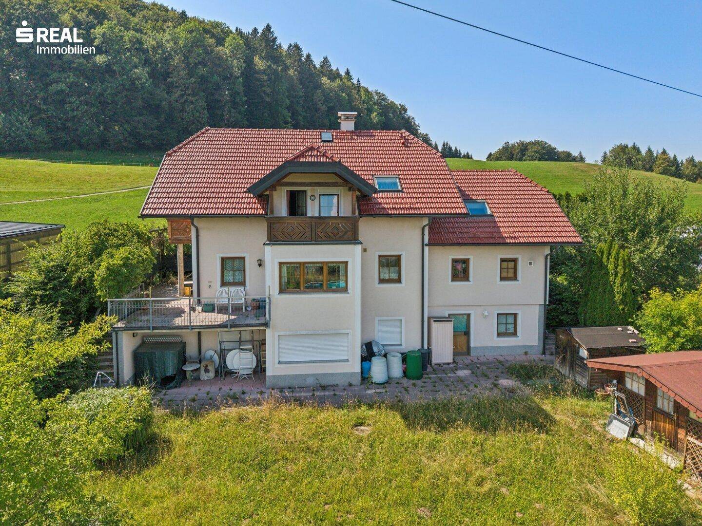 Rückansicht des Hauses mit ungemähter Rasenfläche, Terrasse und Balkon, umgeben von Natur.