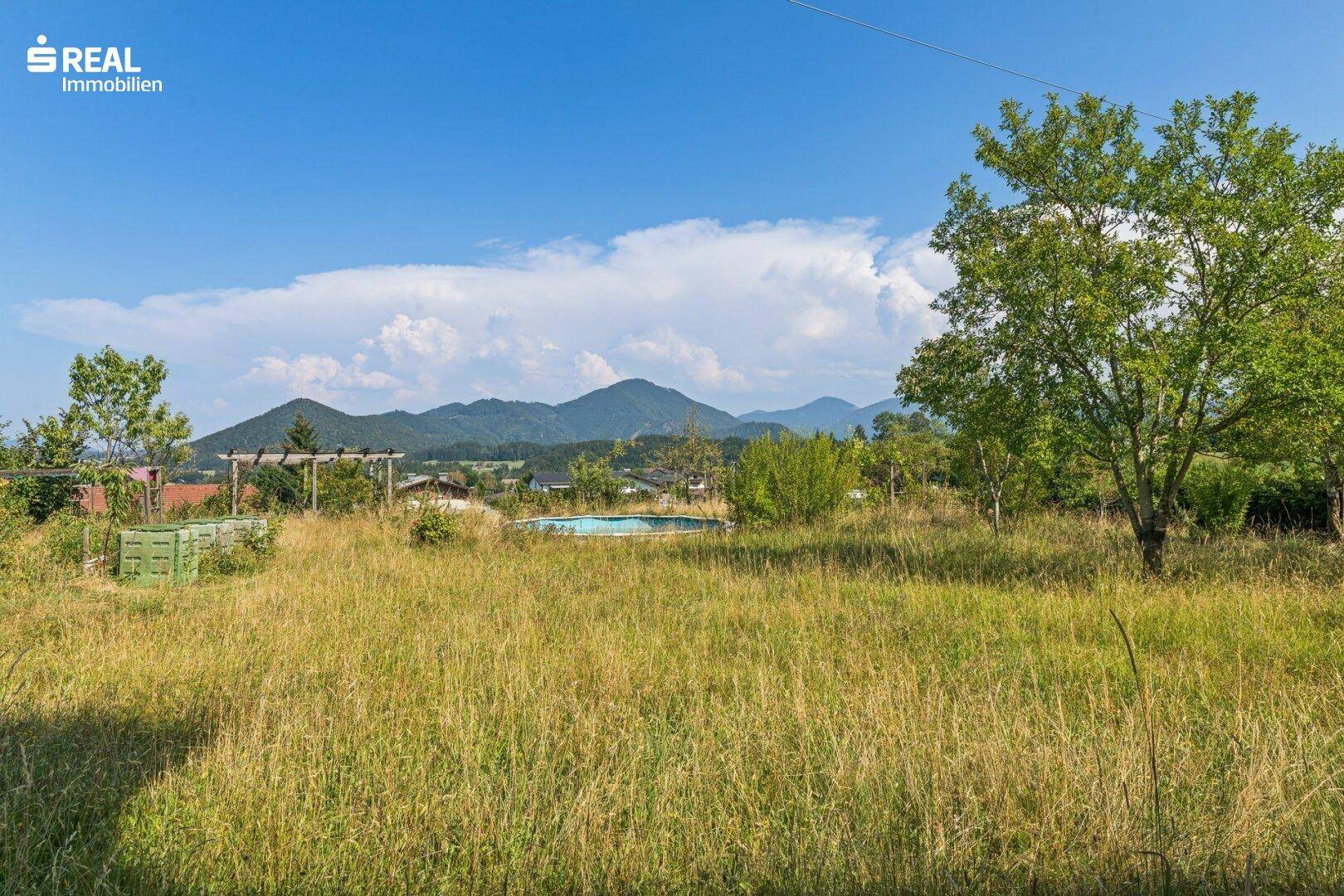 Weitläufige, ungemähte Rasenfläche mit kleinem Pool und Bergblick in ländlicher Umgebung.