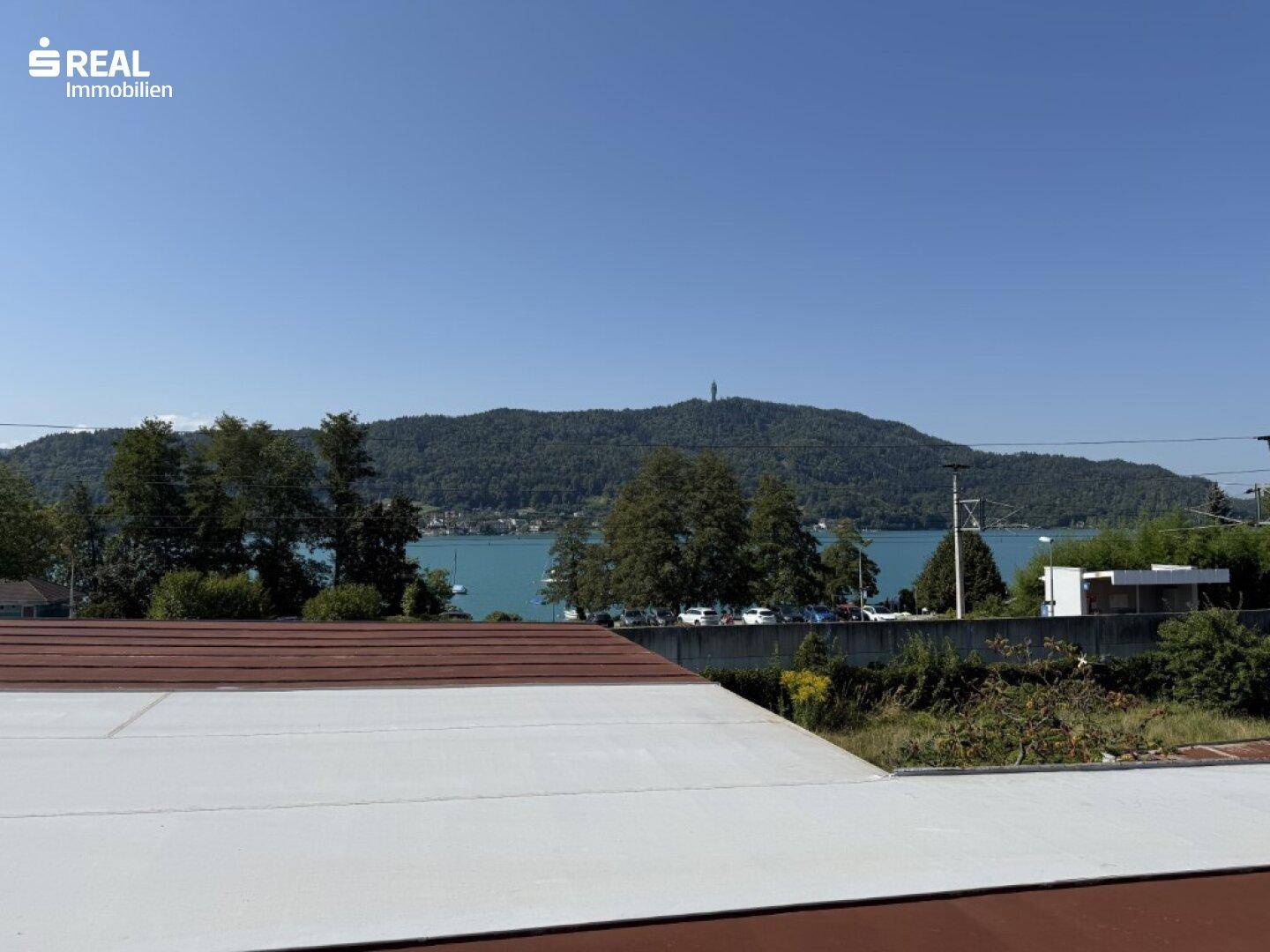 Weitblick über ein Dach auf einen See und bewaldete Berge unter klarem Himmel.