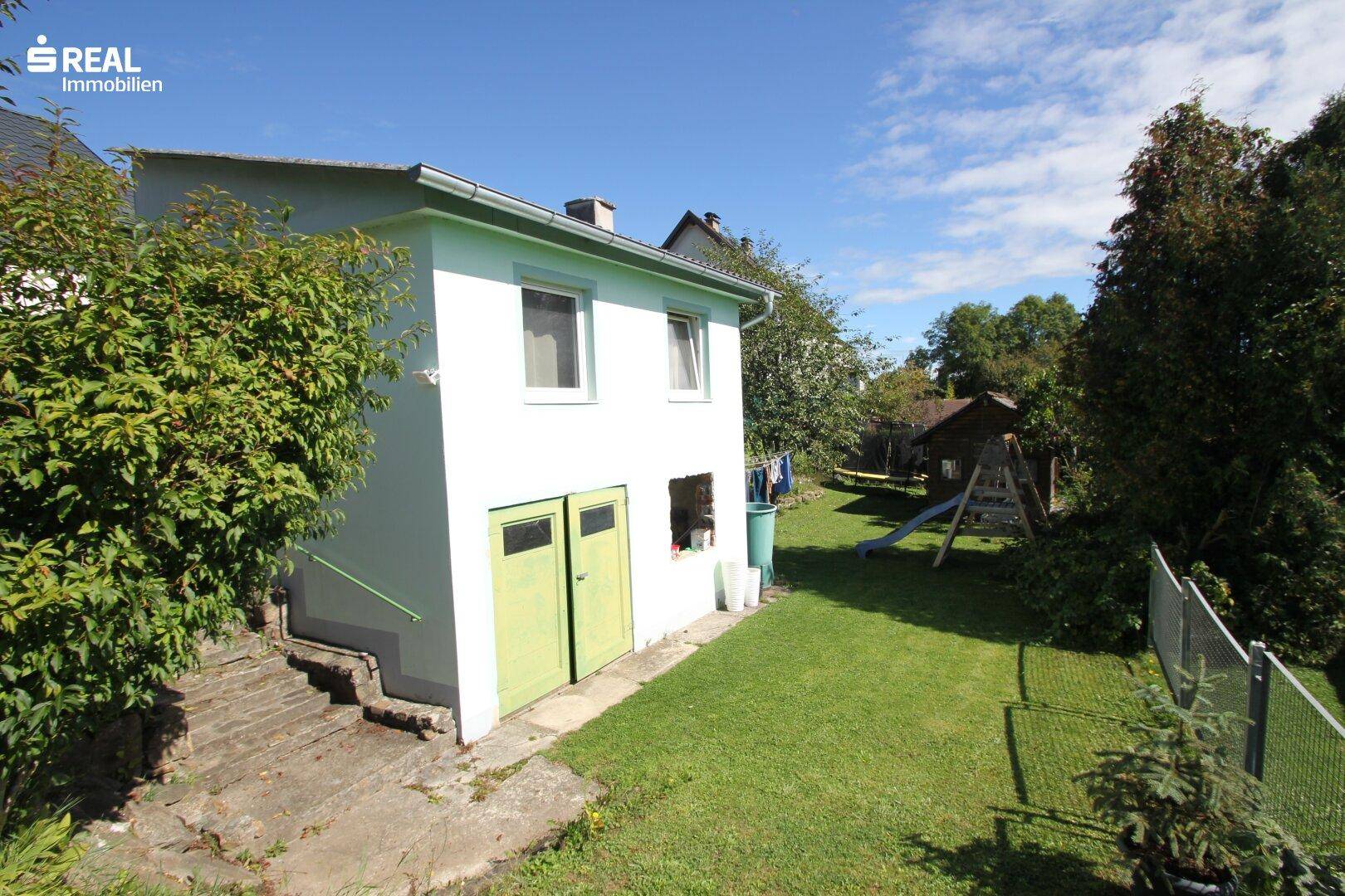 Hinteransicht des Hauses mit grünem Anbau, Garten und Spielbereich im Freien.
