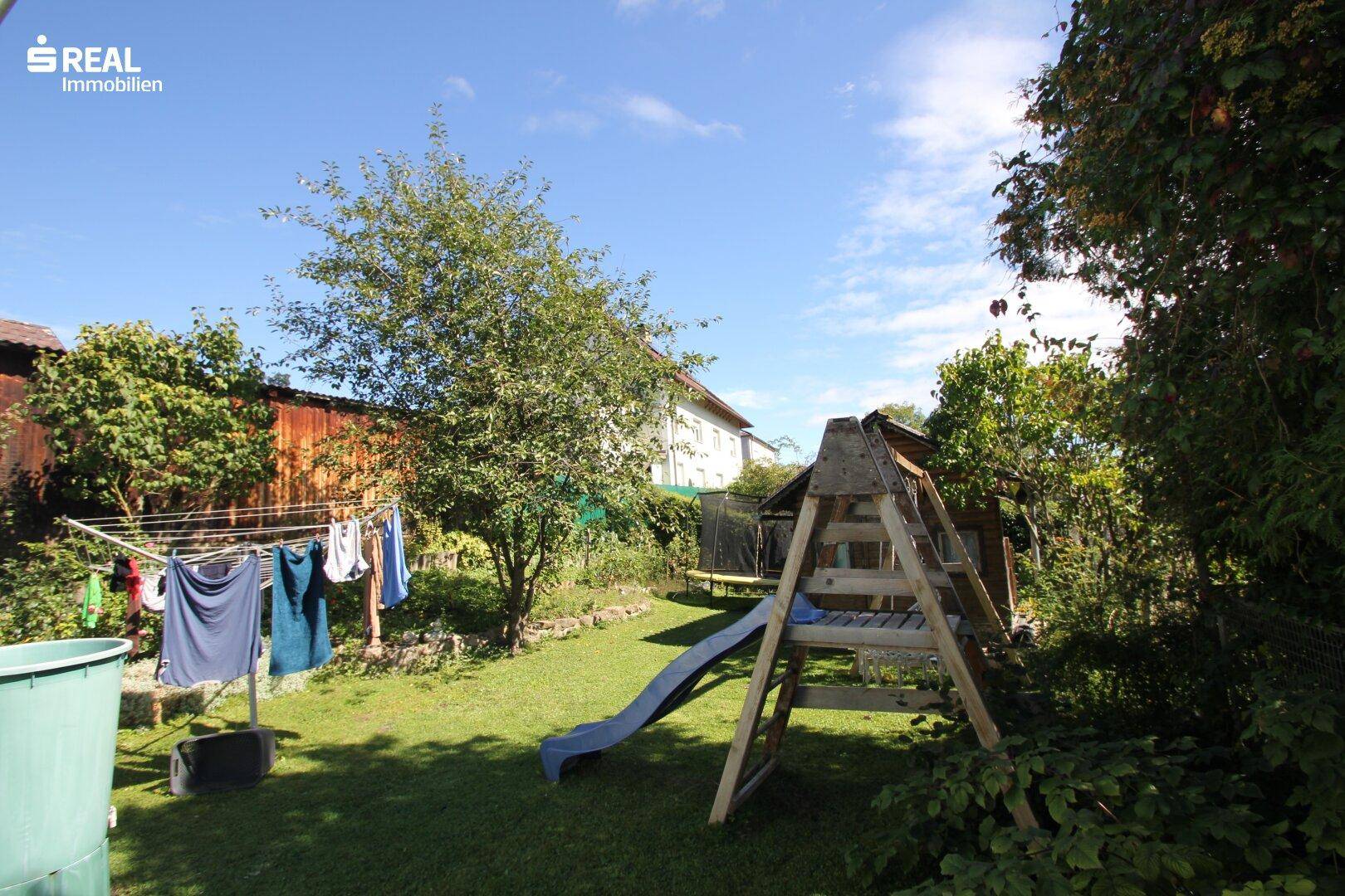 Grüner Gartenbereich mit Rasenfläche, Spielgeräten und Wäscheleine unter sonnigem Himmel.