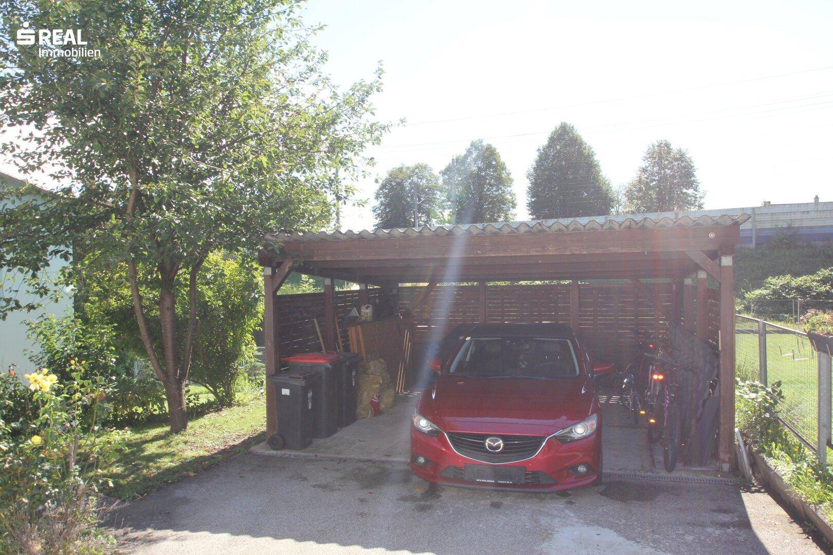 Überdachter Carport-Bereich mit einem geparkten Auto und umgebender Bepflanzung.