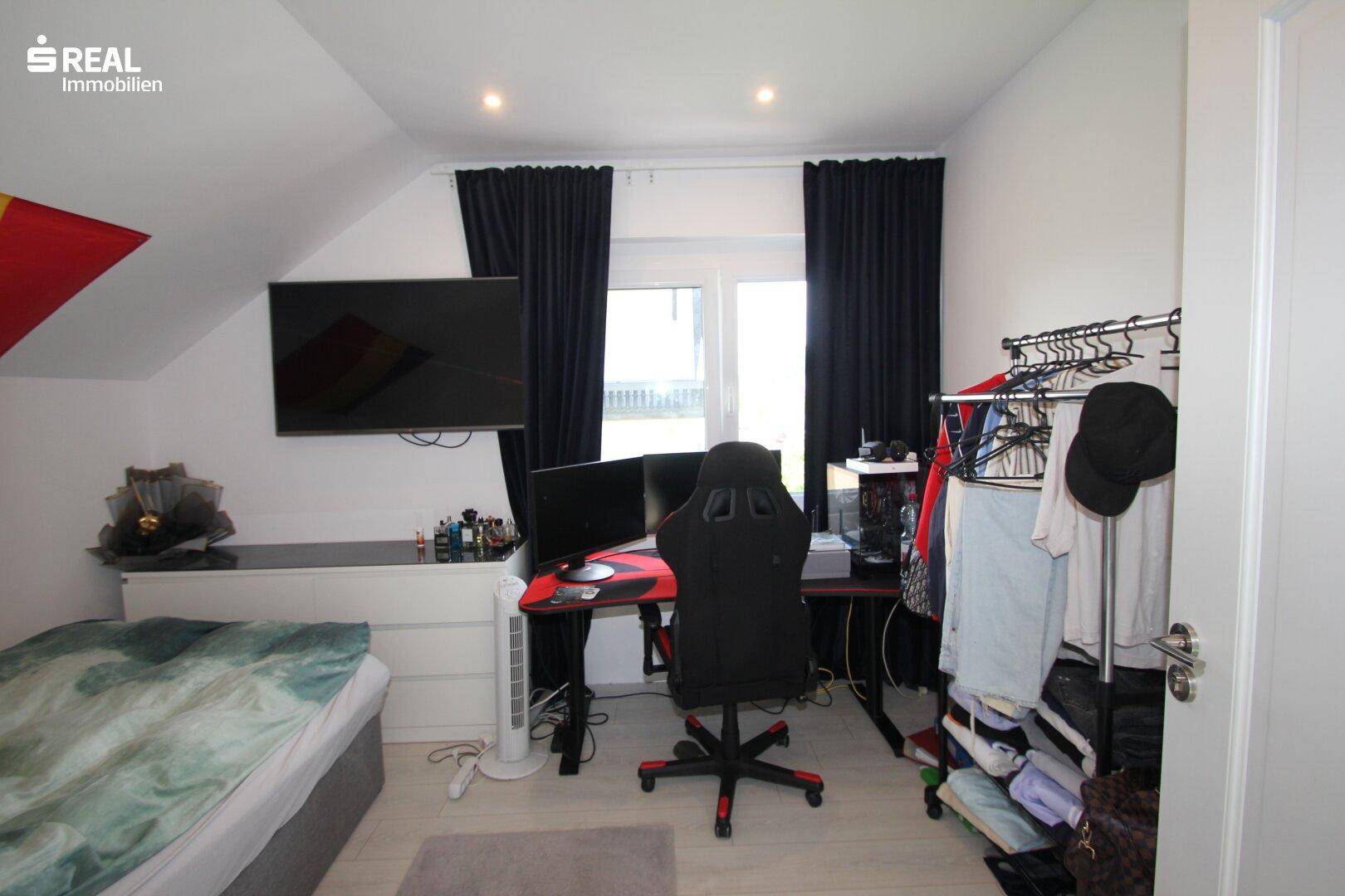 Schlafzimmer mit Dachschräge, Schreibtisch, Gaming-Setup und dunklen Vorhängen.