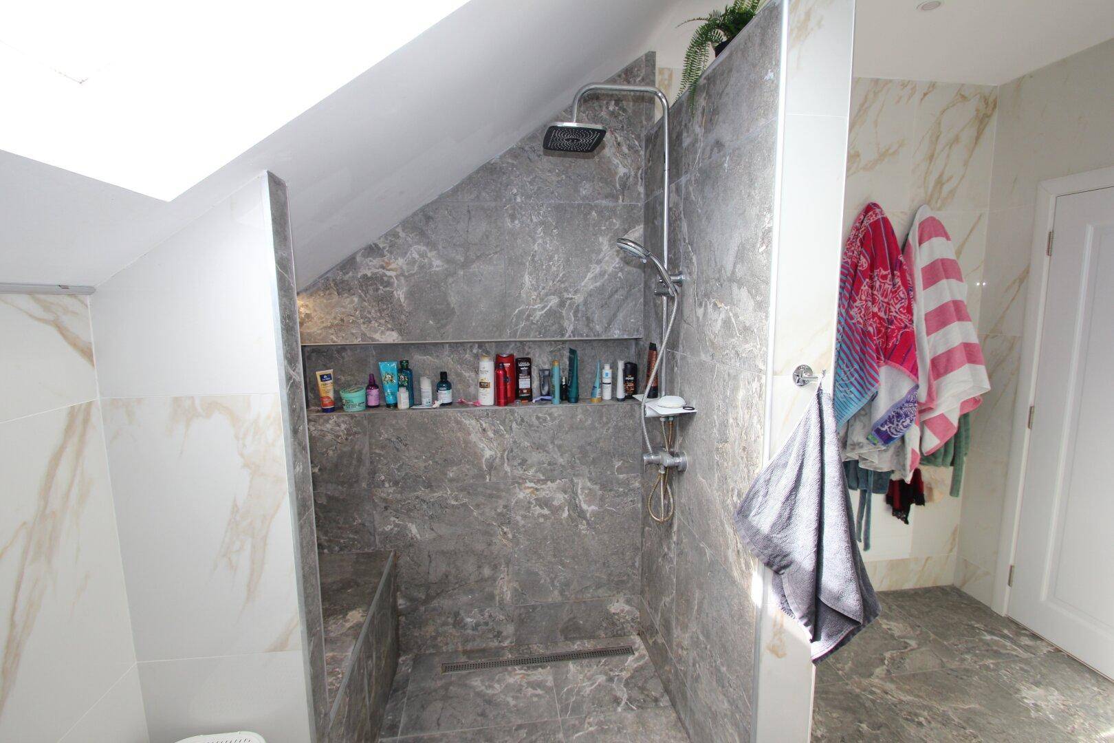Geräumige begehbare Dusche mit dunklen Fliesen und integrierten Ablagen unter einem Dachfenster.