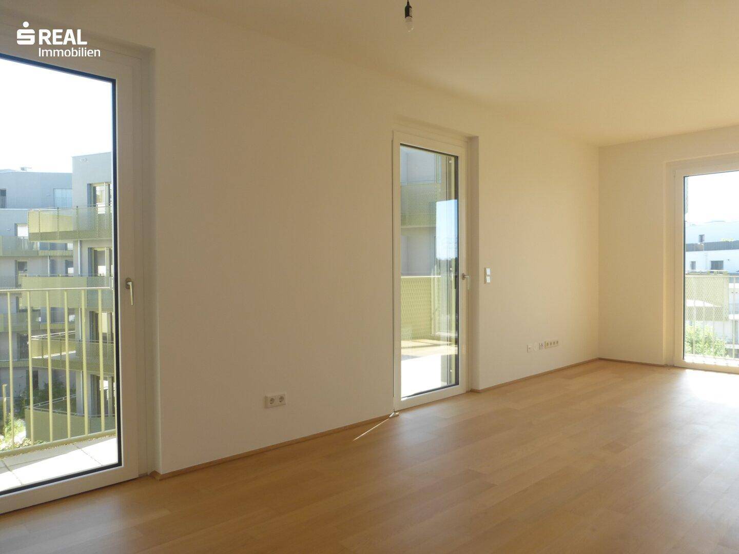 Helles, leeres Wohnzimmer mit Parkettboden und Zugang zu mehreren Balkonen in einem Neubau.