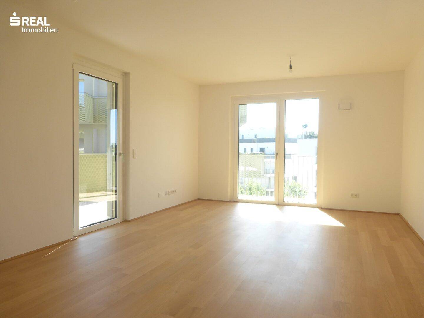 Lichtdurchflutetes Wohnzimmer mit Holzboden und großen Fenstern, die viel Sonnenlicht hereinlassen.