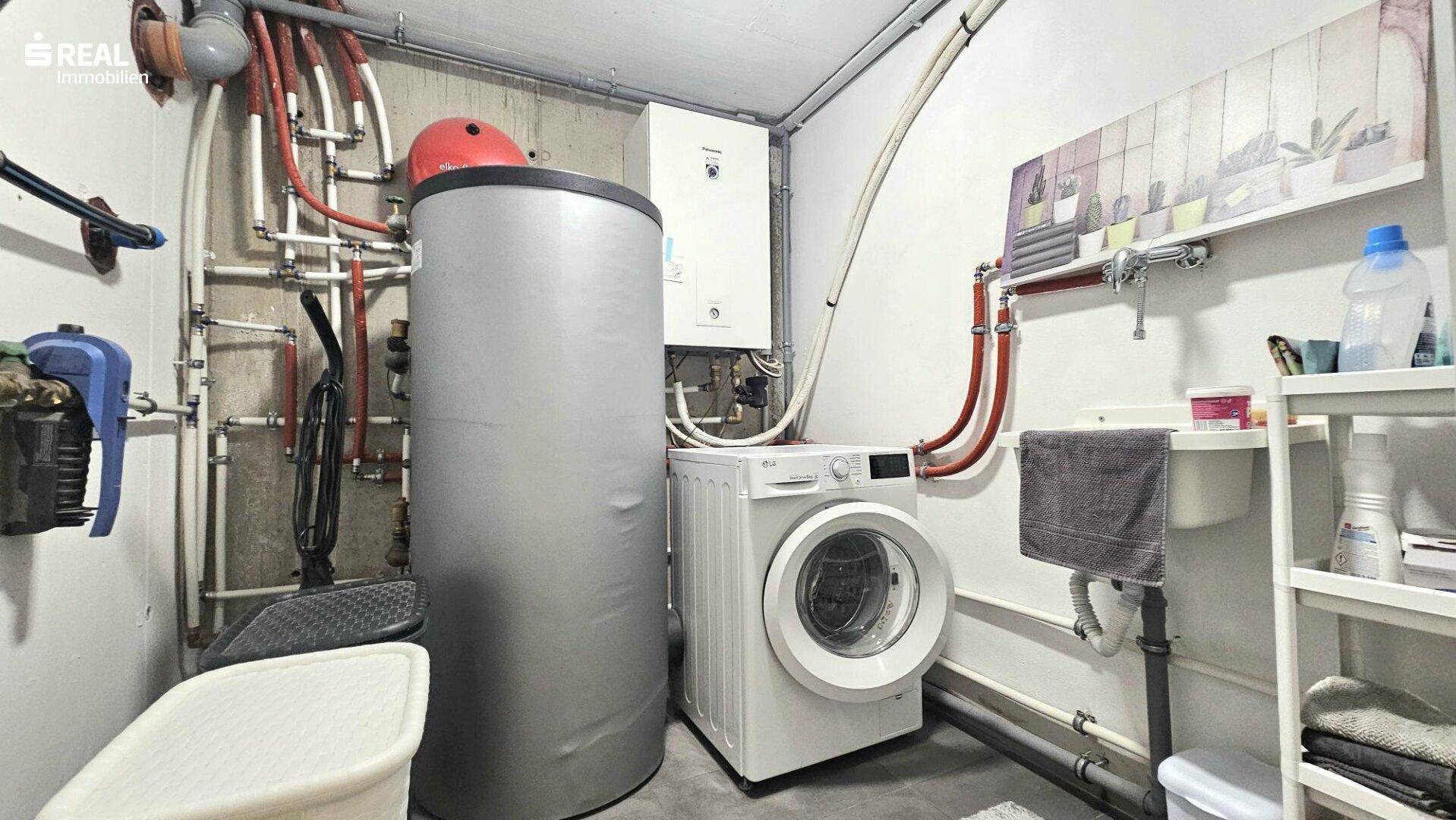 Hauswirtschaftsraum mit Waschmaschine, Warmwasserspeicher und sichtbaren Installationen.