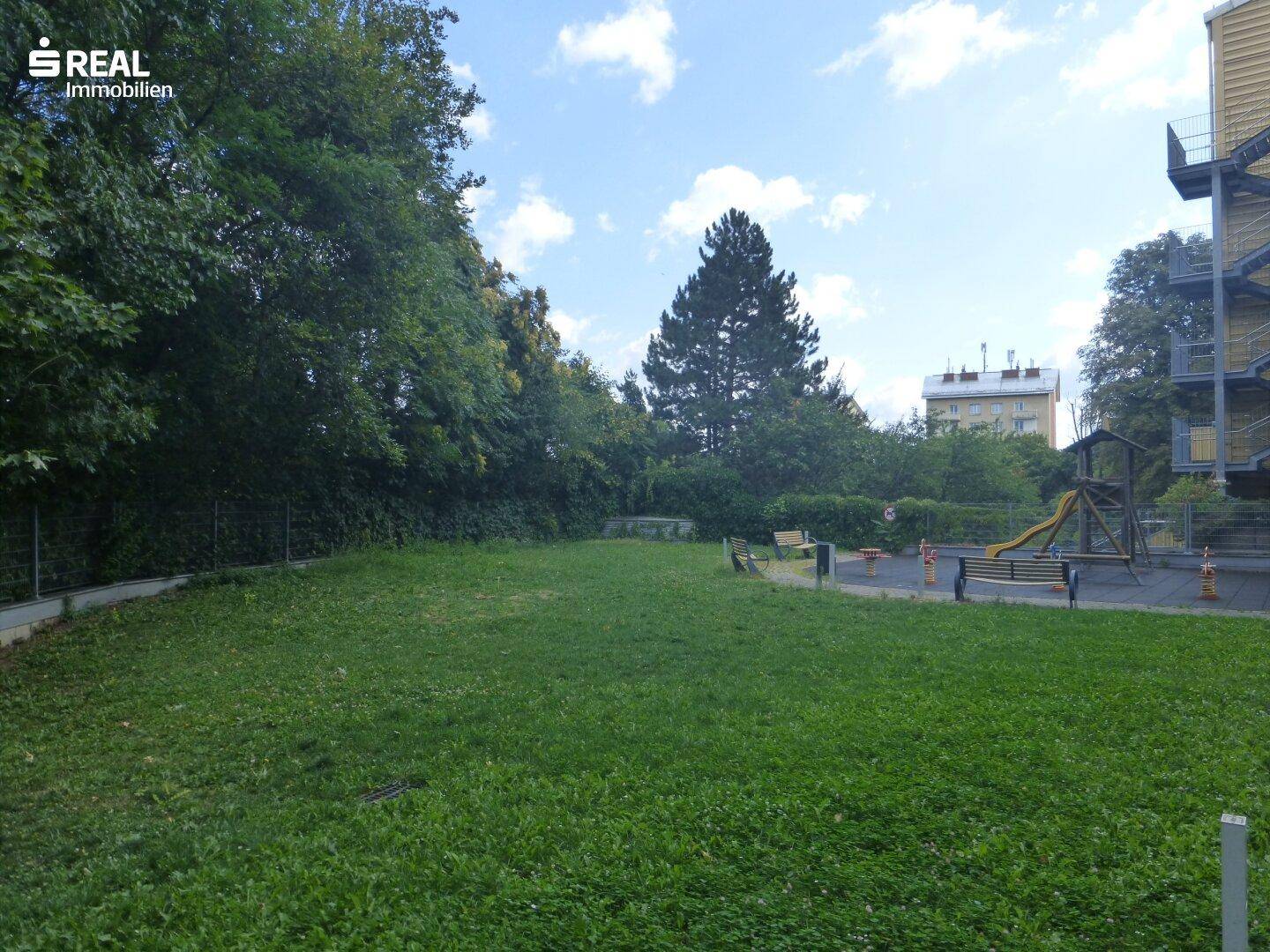 Große Rasenfläche mit Bäumen und einem Spielplatz im Hintergrund.