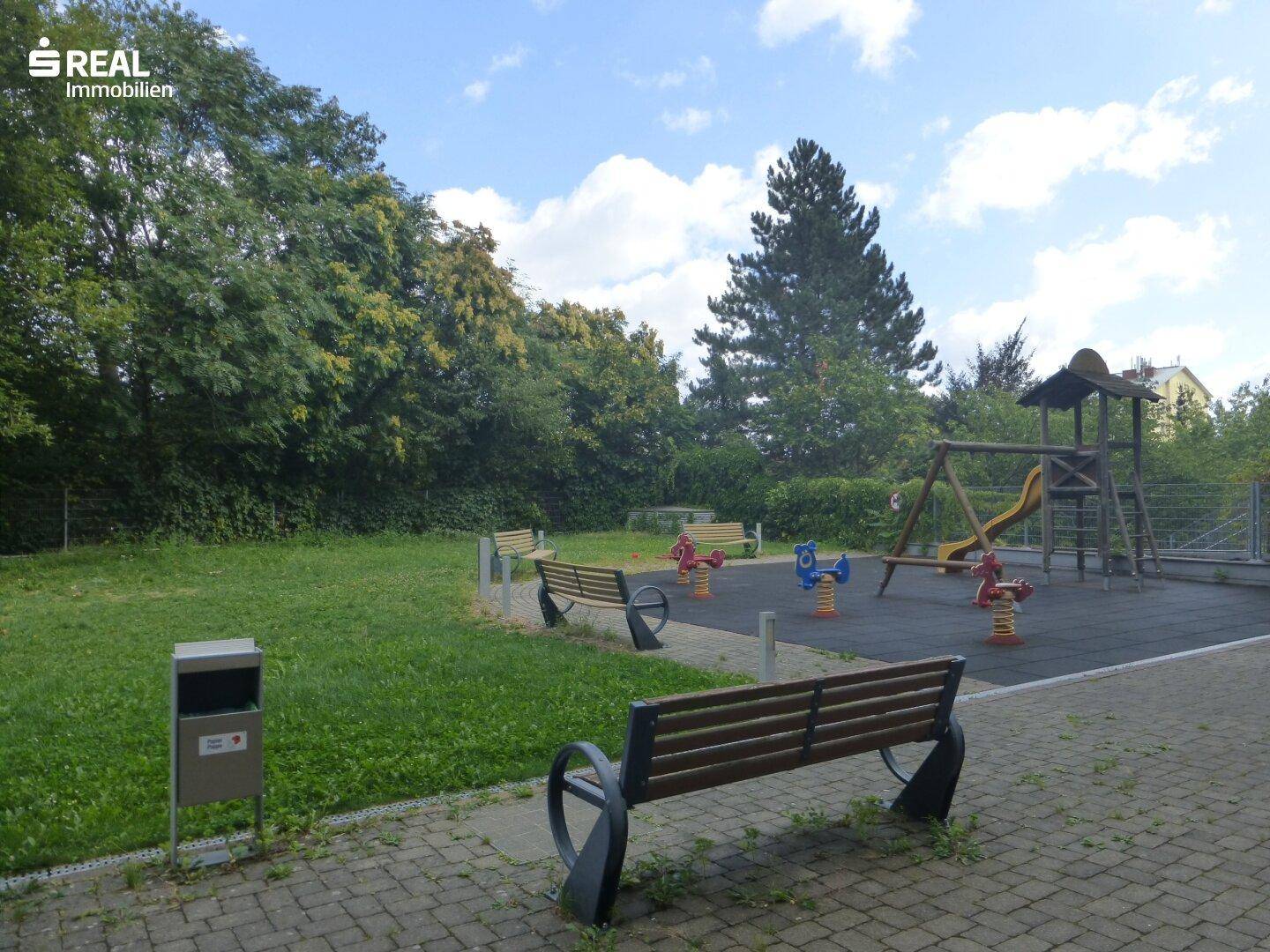Gemeinschaftlicher Spielplatz mit Bänken, Rasenfläche und verschiedenen Spielgeräten.