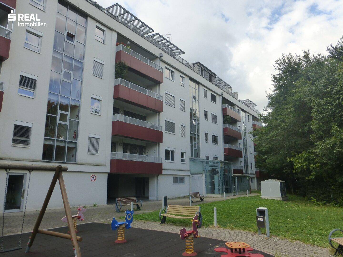 Modernes Mehrfamilienhaus mit Balkonen, Glasfassade und einem Spielplatz im Vordergrund.