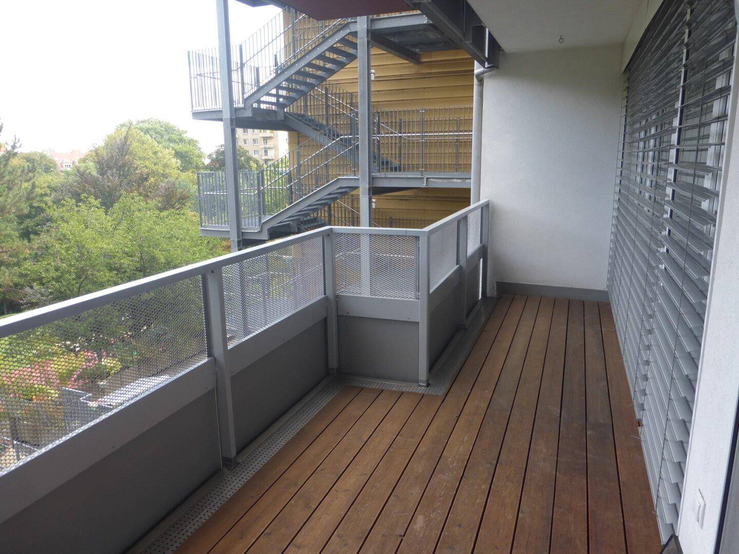 Balkon mit Holzboden und Blick auf Bäume sowie eine externe Metalltreppe.