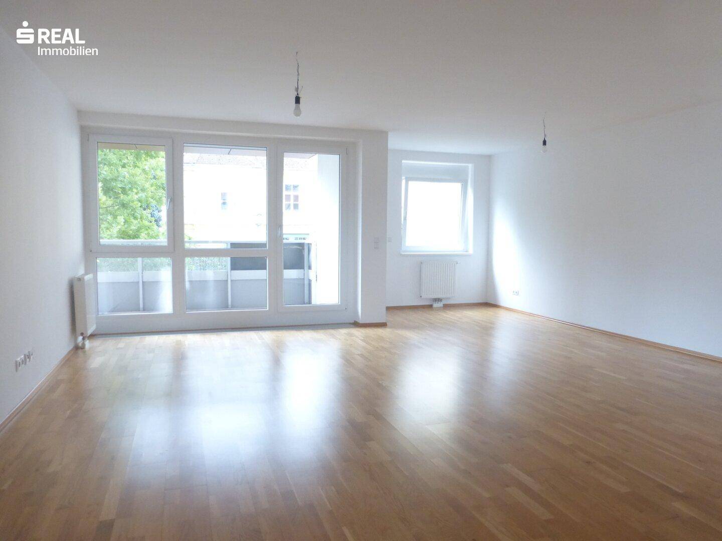 Heller Wohnraum mit Parkettboden, großen Fenstern und Zugang zu einem Balkon.