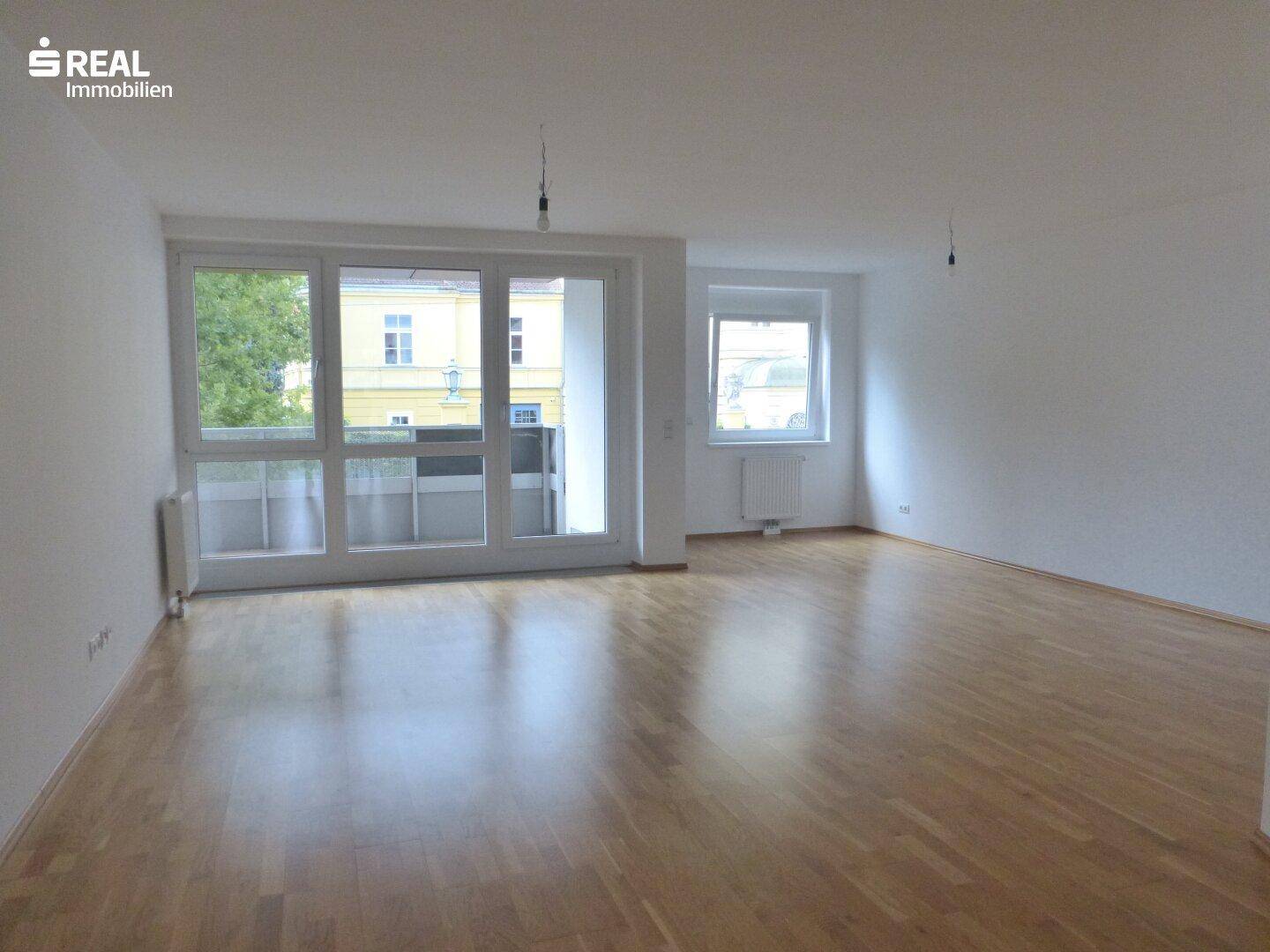 Heller Wohnraum mit Parkettboden, großen Fenstern und Zugang zu einem Balkon.