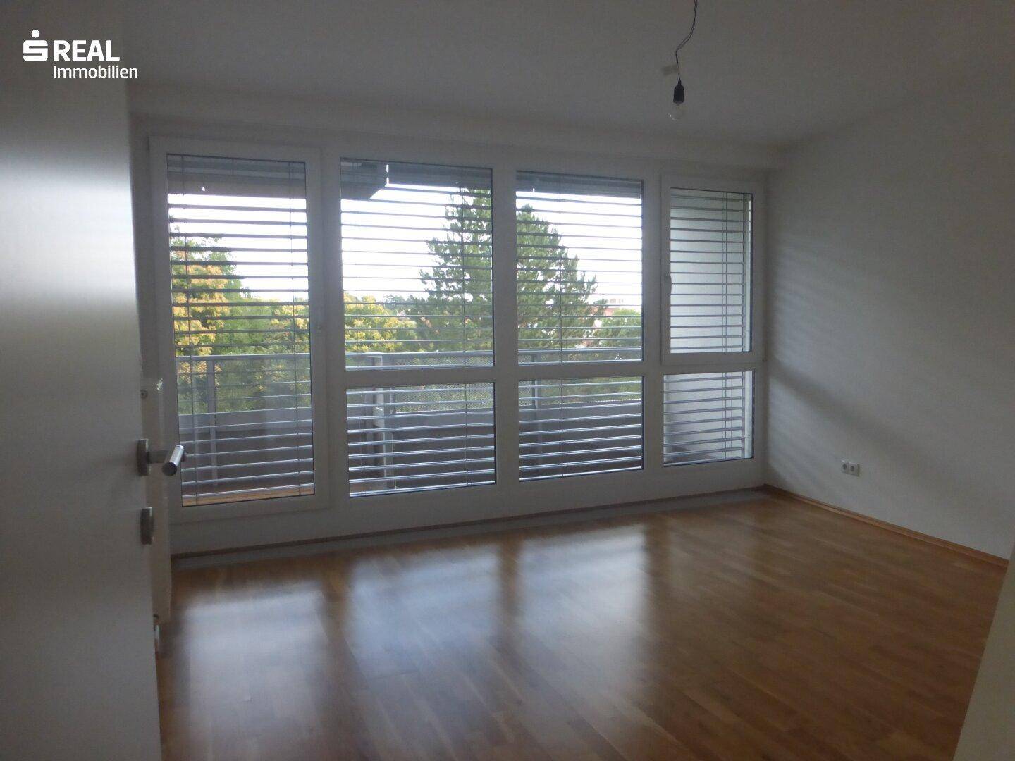 Helles Schlafzimmer mit Parkettboden und großen Fenstern mit Jalousien.