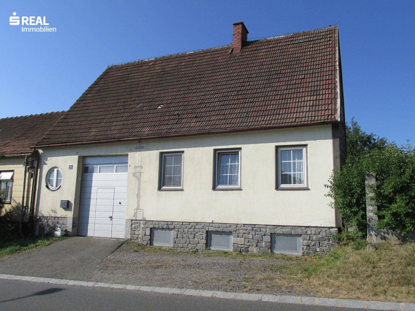 Einfamilienhaus von vorne mit Garagentor, hellbeiger Fassade und Ziegeldach an einem sonnigen Tag.