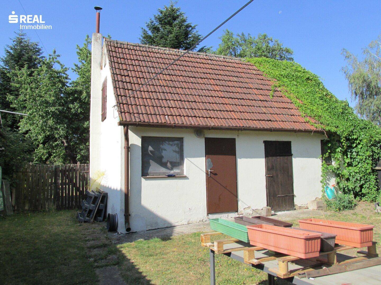Kleines Nebengebäude im Garten mit Ziegeldach, weißer Fassade und zwei Holztüren.