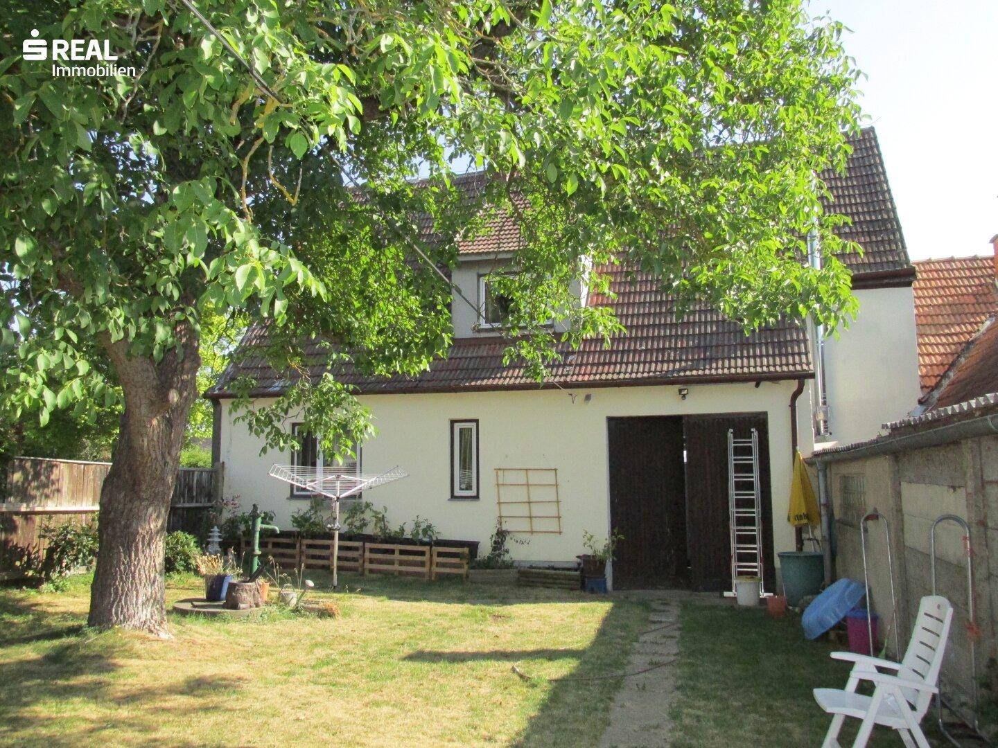 Hinteransicht des Hauses mit großem Baum, Rasenfläche und Zugang zu einem Nebengebäude.