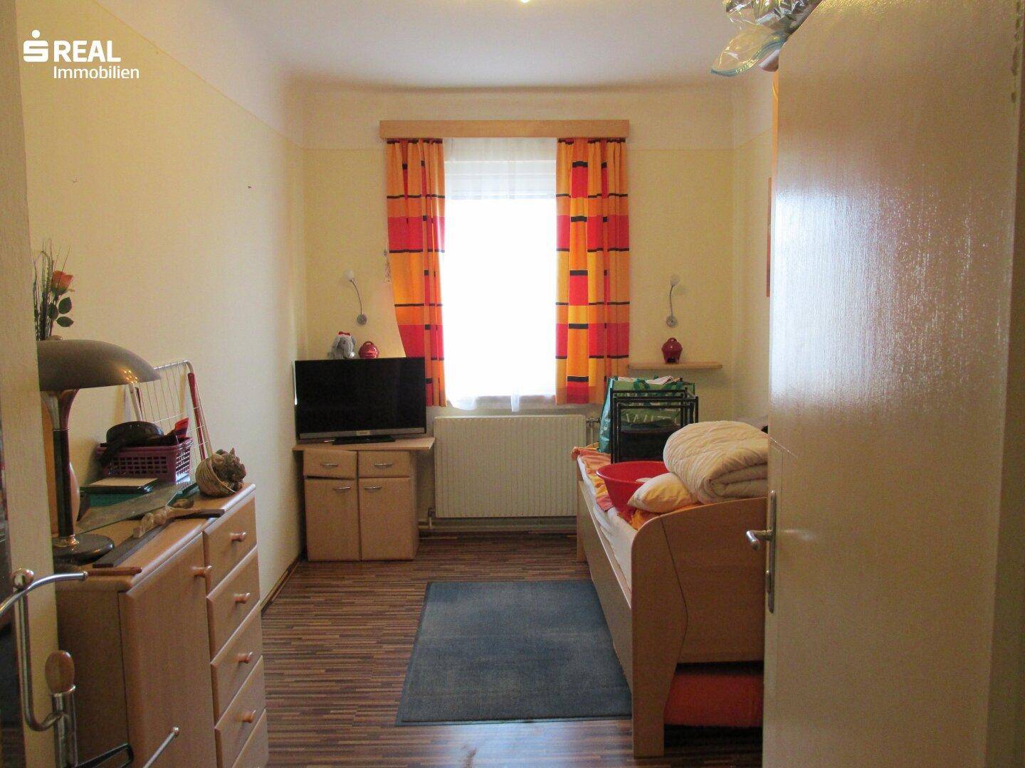 Schlafzimmer mit Einzelbett, Kommode und Fenster mit bunten Vorhängen.