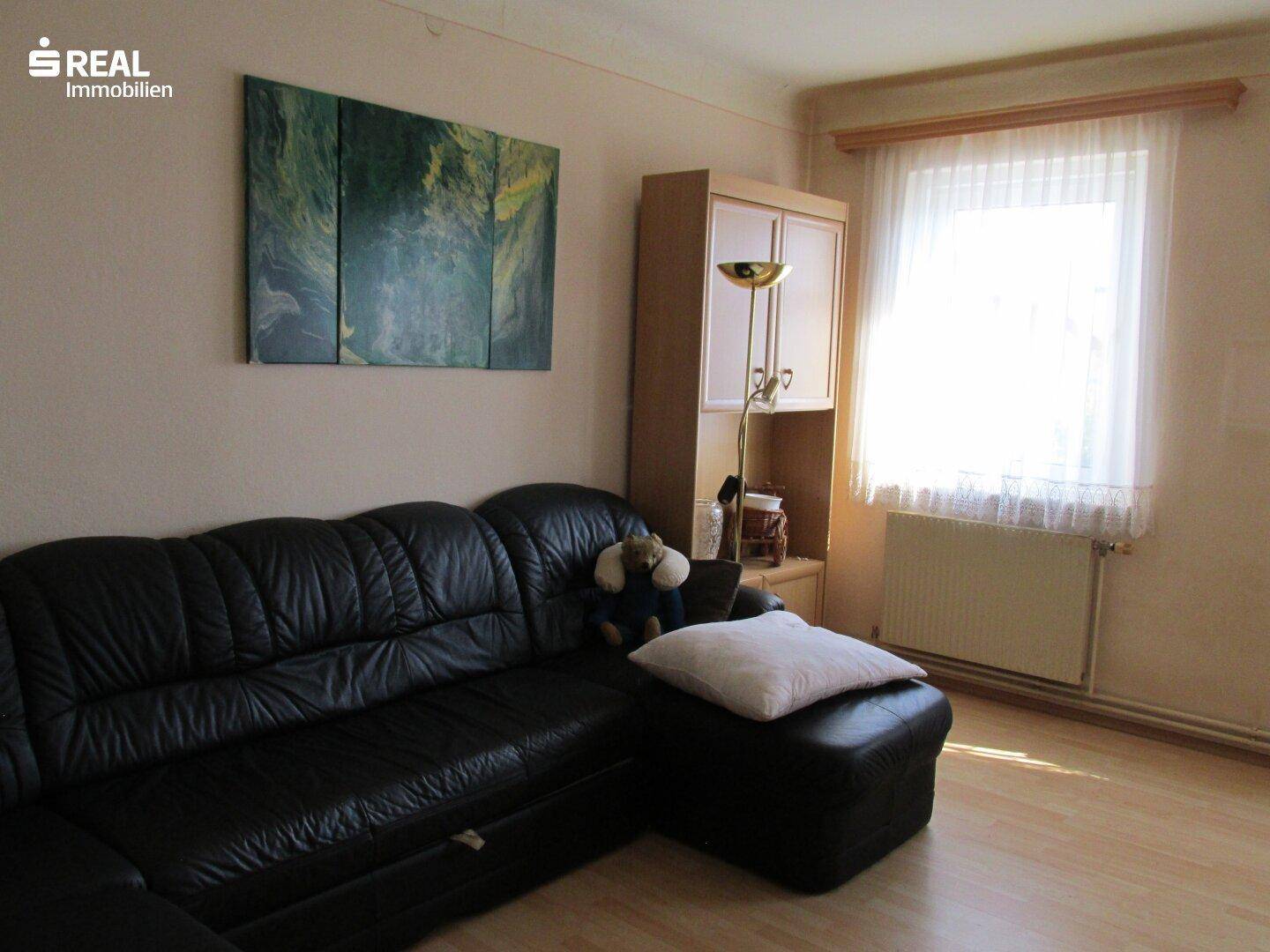 Wohnzimmer mit schwarzem Ledersofa, hellem Laminatboden und Fenster mit Tageslicht.