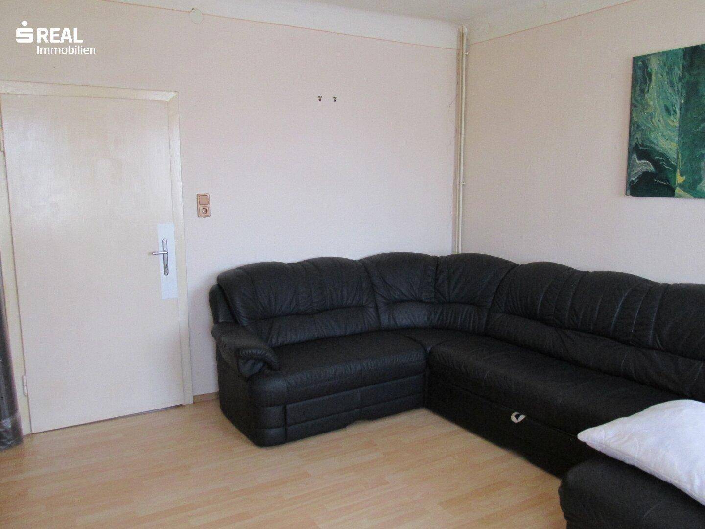 Wohnzimmer mit großem schwarzem Ledersofa, hellem Laminatboden und weißer Tür.